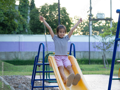 Cheerful little Asian girl raising arms and sliding down yellow slide at outdoor playground
