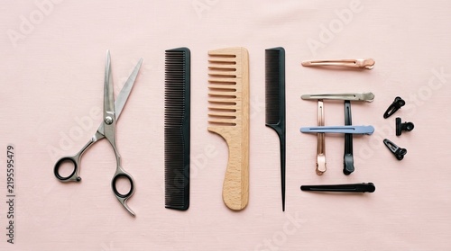 A collection of hair styling tools rests on a pink surface. A pair of scissors, a comb, a wide-tooth comb, and several hair clips are arranged in a linear fashion.