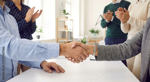 Business handshake over white desk with coworkers clapping in bright workspace.