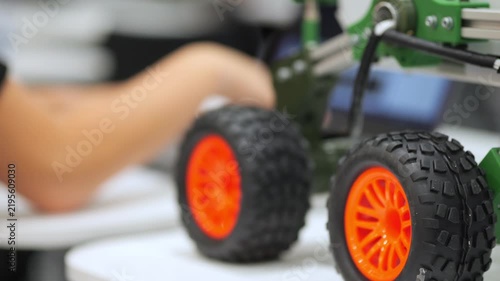 Close-up of orange wheel toy car. Blurred hands work nearby. Green robotic vehicle detail. Tech workshop environment shown