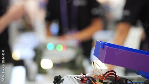 Close-up of electronic circuit board with wires and glowing lights. Background shows blurred people at tech event. Focus on technical components and active devices