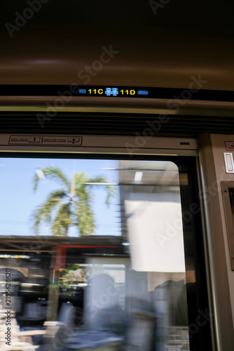 Train interior viewed from a moving passenger seat. Digital sign displays station numbers above the window. Palm trees blur past as train glides through sunny landscape. Passengers' forms indistinct
