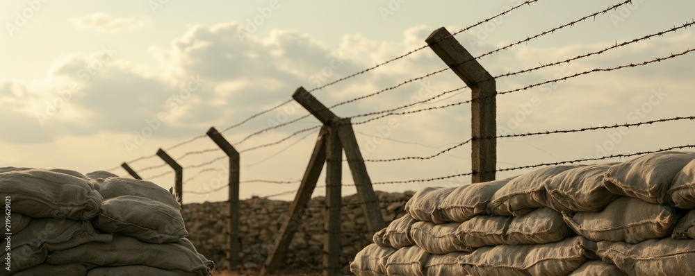 Obraz premium A barbed wire fence stretches alongside sandbags under a cloudy sky, suggesting a military or conflict zone setting at dusk.