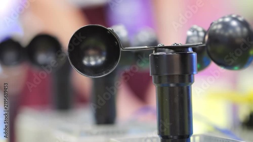 Close-up of black anemometer with rotating cups. Multiple similar instruments visible in background. Blurred indoor setting suggests science fair or exhibition
