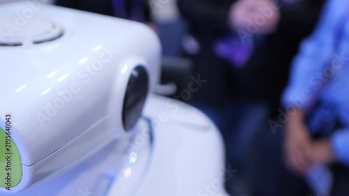 Close-up of white robotic head with camera lens. Blurred background shows people at event. Robot has green light and circular sensor. Focus on robot's facial features