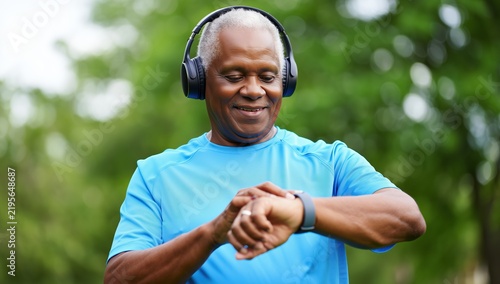 African American senior man checking smartwatch during outdoor workout wearing wireless headphones and blue athletic shirt in park setting with green foliage
