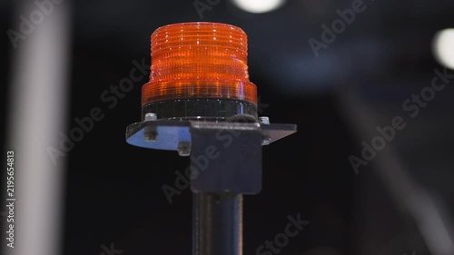Orange warning light glowing steadily on metal pole. Close-up shot shows bright illumination. Dark background highlights the light's intensity. Light appears in multiple frames, steady and focused