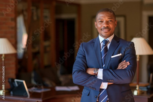 Confident businessman in a dark blue suit standing with arms crossed inside an office room