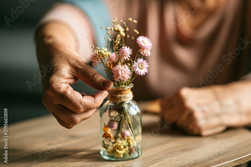 Elderly woman hands arranging small dried flowers in a glass bottle on wooden table