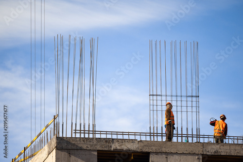 Two workers stand atop unfinished concrete building under blue sky