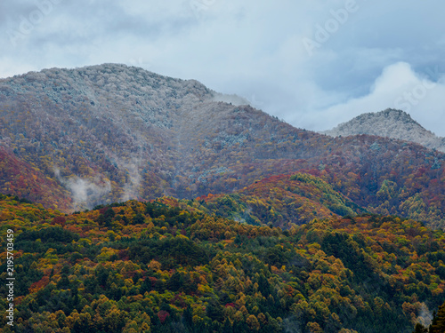 Фотография 晩秋の風景。秋と冬の季節が重なる様子。霧氷した山頂、中腹の紅葉、麓の緑の樹木の色彩の変化。グラデーション。三段紅葉。自然風景素材。美しい志賀高原。