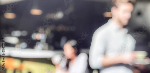 A blurred image of people in a café setting. One person is holding a drink while another sits in the background. The atmosphere suggests a casual social environment.