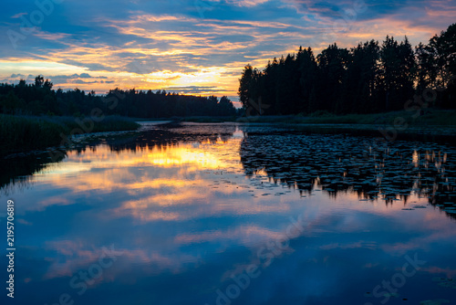 A magical landscape featuring a beautiful sunset or sunrise over a pond or lake on a summer evening, with a cloudy sky in the background, reflections on the water
