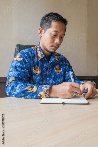 A concentrated man in a blue traditional batik shirt writes notes in a notebook at an office desk, conveying focus, planning, professionalism and everyday work routine.