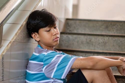 A depressed teenage boy sits on school stairs with his eyes closed, leaning against a tiled wall.