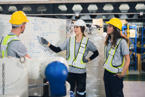 Factory team discusses technical diagrams on a whiteboard, with one worker production details during an industrial engineering planning meeting