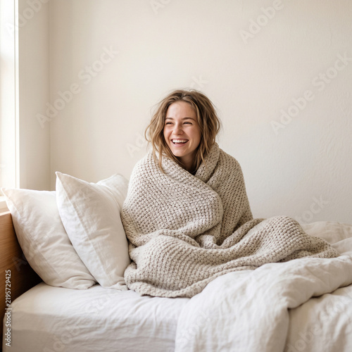 Happy young woman wrapped in cozy blanket laughing in bed, cheerful female relaxing during comfortable morning routine, concept of home comfort, wellness, and mental health