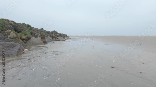 Cinematic low-angle shot moving along a misty and desolate beach with a rocky breakwater on an overcast day.