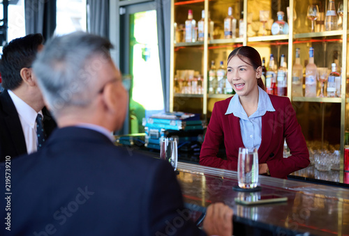 A smiling female bartender in a red blazer assists a male customer at a polished bar, with bottles of spirits and colorful drinks visible on shelves in the background