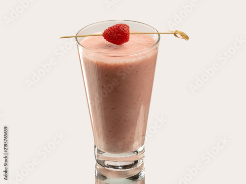 Tall glass of pink strawberry smoothie topped with a fresh strawberry on a skewer, shot on a light background.