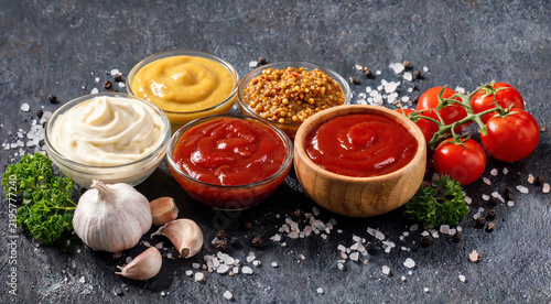 Different types of sauces in bowls with fresh ingredients on a dark surface