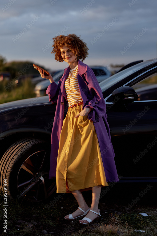 Fototapeta premium Young woman with curly hair walks beside a dark car, wearing a purple coat, striped top, and yellow skirt, expressing a vibrant outdoor lifestyle and confident casual fashion.