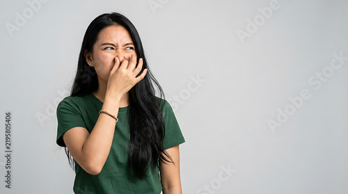 An Asian woman pinches her nose with her fingers to block a foul stench while expressing extreme disgust and disapproval due to a terrible smell drifting through the air around her room