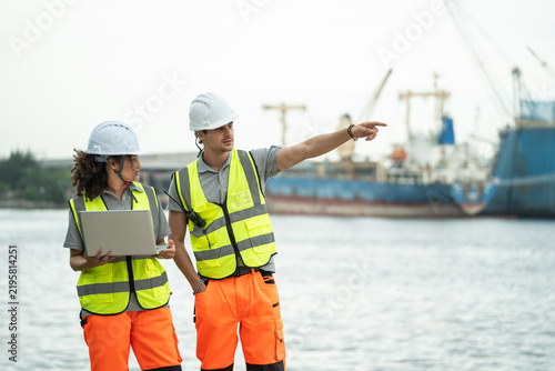 Obraz na płótnie Port engineers wearing safety vests and hardhats stand by the harbor while one points forward and holds a laptop, communicating about ongoing freight logistics and operations at the cargo ship