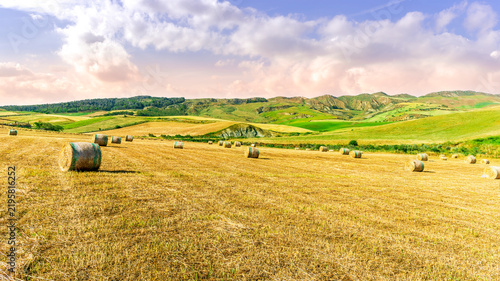 Scenic view at picturesque golden sunset valley in a green shiny field with hay stacks, bright cloudy sky , golden sun rays and beautiful hills on background, summer valley landscape