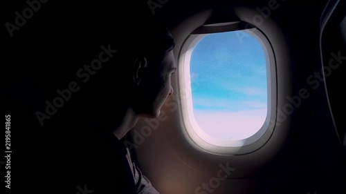 Silhouette passenger gazing through airplane window at blue sky and distant clouds during flight. Young man looking outside aircraft porthole in dark cabin contemplative mood. Air travel view serenity