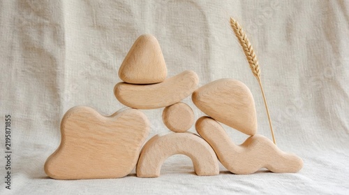 A collection of smooth unvarnished wooden building blocks in organic rounded shapes stacked together next to a dried wheat stalk on a neutral beige linen fabric background.