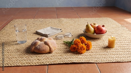 Traditional pan de muerto and cempasuchil flowers. Day of the Dead altar setup on woven straw mat. Fresh fruit bowl, water glass, and lit candle