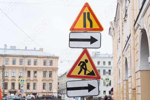 Yellow triangle road signs indicating construction work and road narrowing ahead. Group of traffic signs with directional arrow on street post against blurred historic building background.