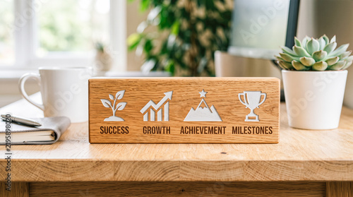 success growth achievement milestones Engraved oak wood block sign displaying business icons and motivational words sitting on bright office desk beside coffee mug