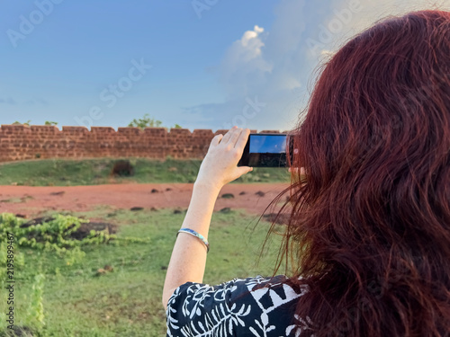 Woman with red hair taking a picture with her smartphone at the ancient Chapora Fort in Goa, India