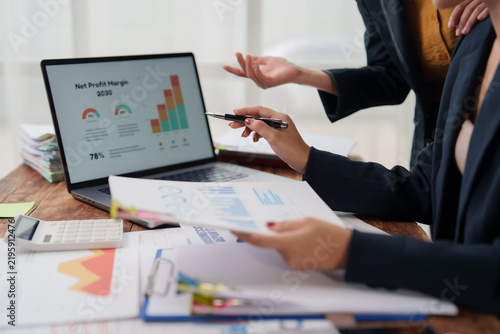 Business professionals collaborating at a desk, reviewing charts and financial reports on a laptop and printouts while discussing data-driven strategy, planning and performance