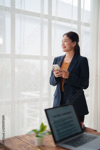 Foto Asian businesswoman stands by a bright office window holding a smartphone, looki