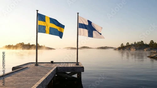 Swedish and finnish flags waving together on wooden pier at sunrise in nordic archipelago sweden finland