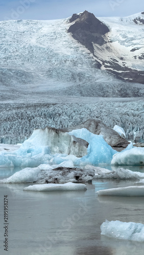 Melting icebergs floating in a glacier lagoon at the foot of an Icelandic glacier, surrounded by cold blue water and rugged mountains.