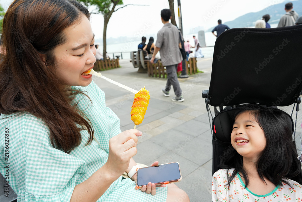 Obraz premium mother pulling melted cheese from a fried corn dog stick while her young daughter sits in a stroller, laughing cheerfully along the scenic Tamsui Old Street boardwalk in New Taipei City, Taiwan.