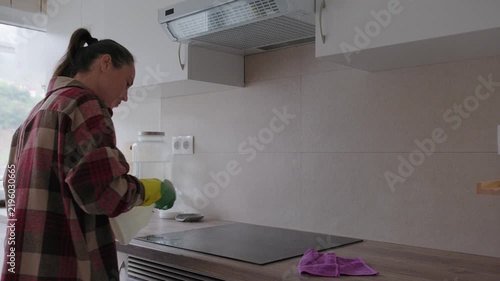 Woman in protective rubber gloves sprays detergent and wipes an induction stove surface with a microfiber cloth in a modern kitchen.