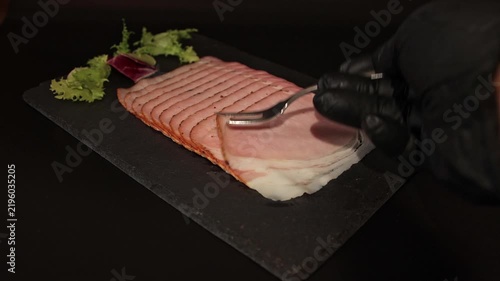 Close-up of chef in black gloves taking a slice of smoked ham with a fork on slate plate. Perfect stock footage for culinary ads, charcuterie videos, and food menus.
