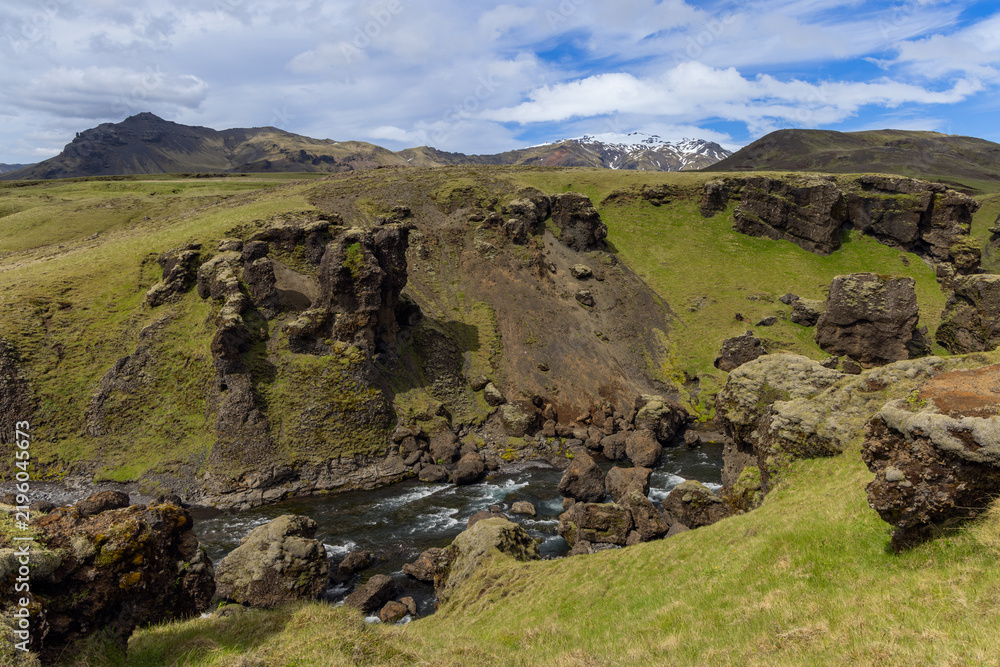 Obraz premium Skoga River flows between dark rock walls and scattered boulders in a grassy Iceland gorge, with rugged ridges and distant snow patches beneath a partly cloudy blue sky.