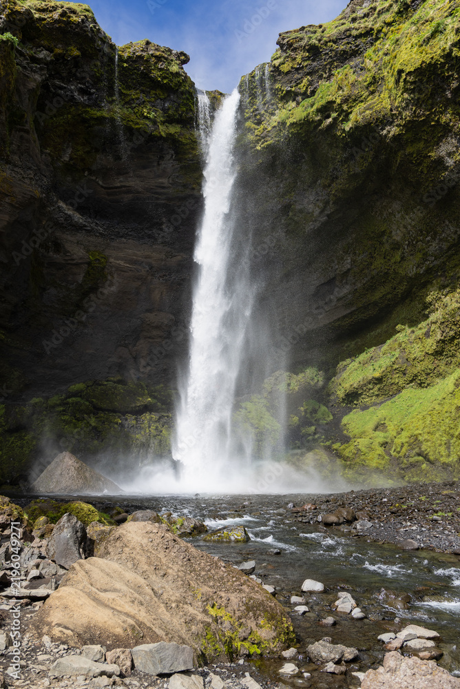 Obraz premium Kvernufoss waterfall drops between moss-covered canyon walls in Iceland, sending spray over a dark pool as the rocky Kverna River flows past a large foreground boulder.