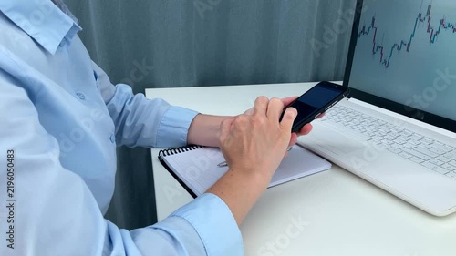 The woman is sitting at a white table, searching for information online, using her phone, assessing market trends using computer charts and records, and planning her further investment strategy