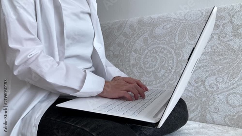 The woman is typing on a laptop, sitting on the sofa, working remotely from home. The concept of digital employment, freelancing, online business, and flexible workspace. Side view