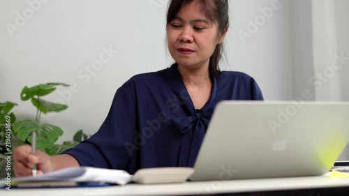 Asian businesswoman looking at laptop screen with thoughtful expression while analyzing financial sales report at desk in bright comfortable corporate office environment room