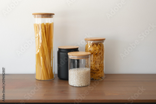 Glass jars filled with spaghetti, rice, and farfalle pasta on wooden kitchen counter. Organized kitchen food storage with modern containers, pantry organization, minimalist home kitchen