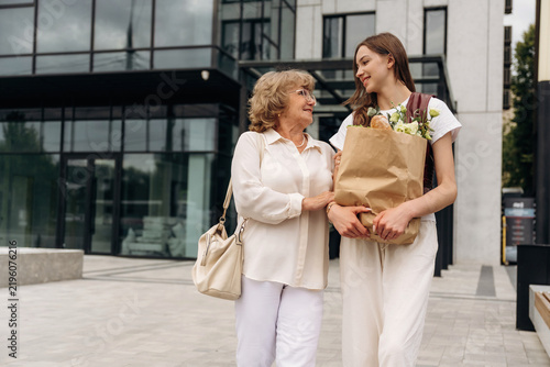 Two women, one elderly and one young, walking together outside a modern building, carrying grocery bags filled with fresh produce and smiling at each other