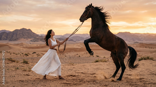 Woman in elegant white dress controlling rearing dark horse on desert dunes at sunset, wild stallion training, taming and freedom concept
